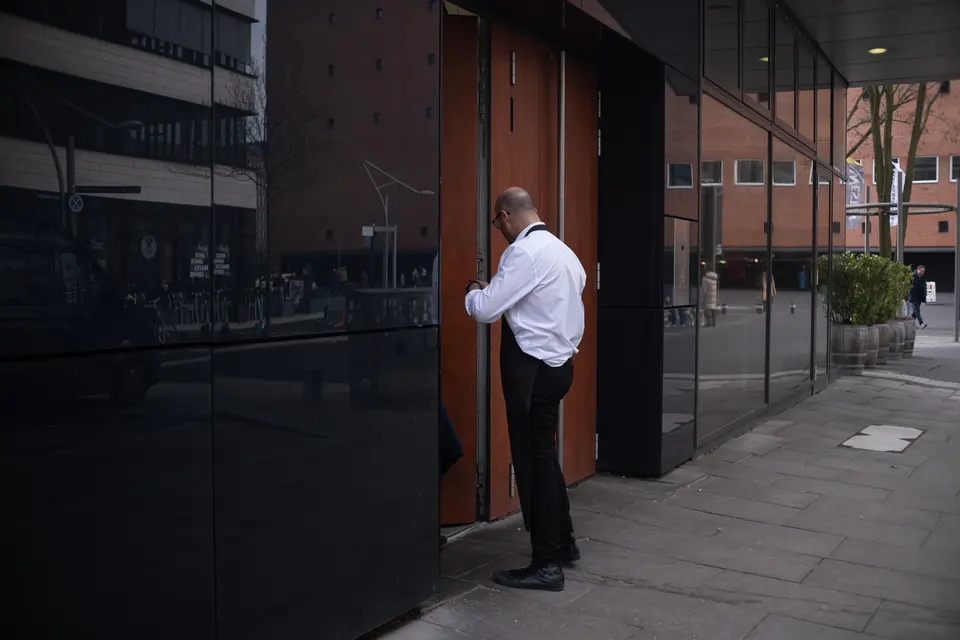 A waiter in a white shirt at a doorway between dark glass façades