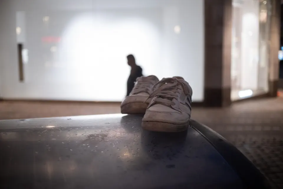 Worn white trainers on a bin at night, a figure in silhouette in a lit window behind