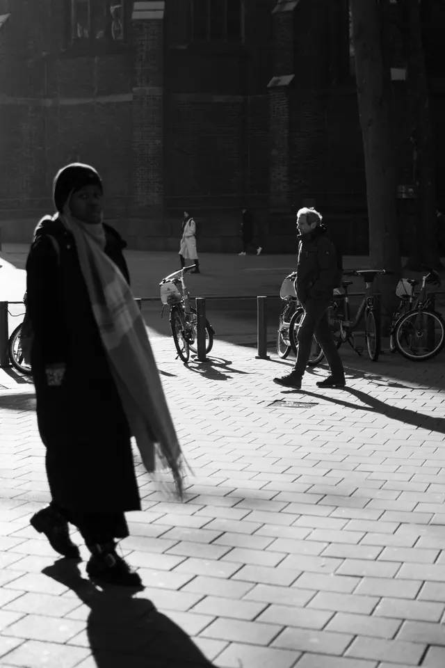 A woman with a long scarf walking through low sunlight, long shadows on the paving, in black and white