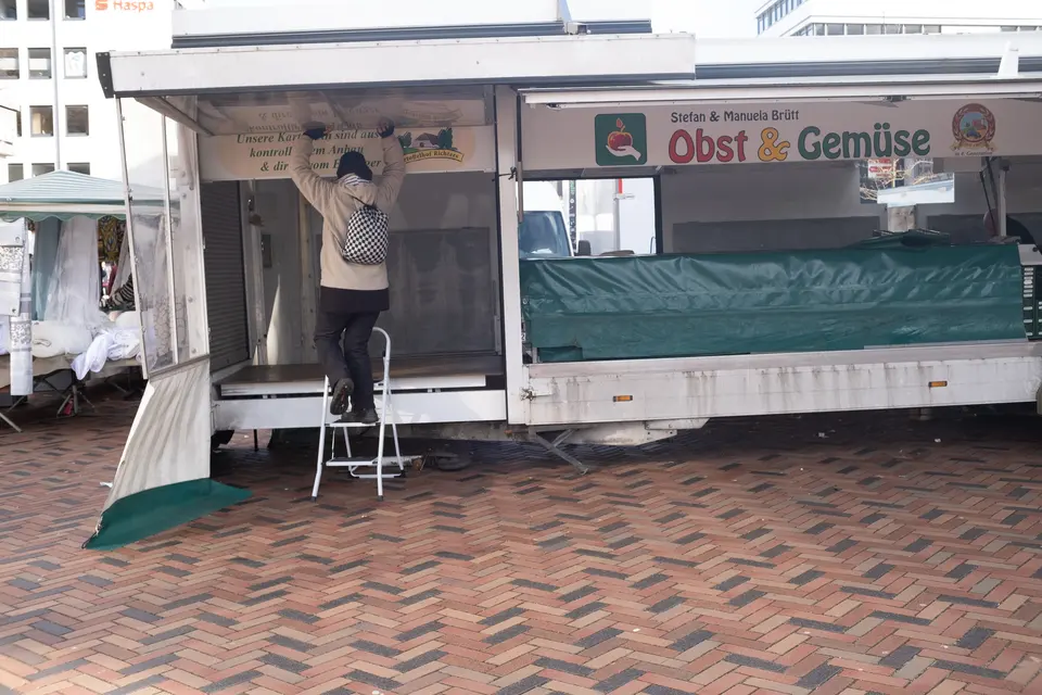 A fruit and vegetable stall being set up at a market, a person on a step ladder at the awning