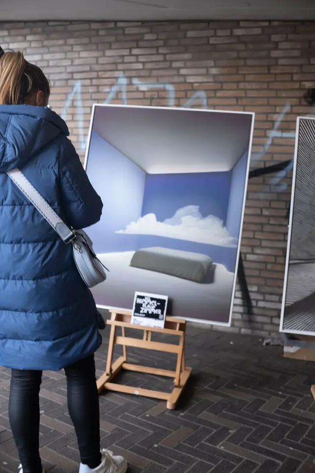 A visitor in a blue coat in front of a painting of a cloud in a room, on an easel outdoors