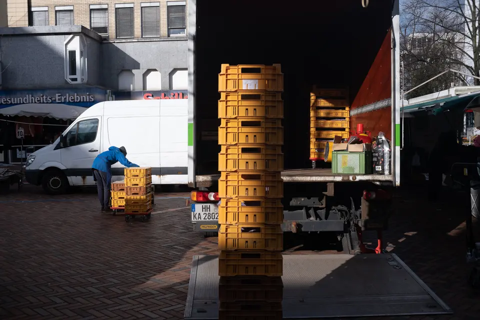 Stacked yellow crates beside a delivery truck at a market, in hard sunlight