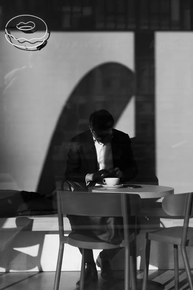 A man at a café table seen through a window, crossed by hard shadows, in black and white