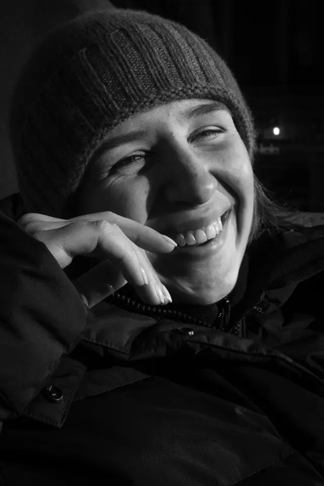 Black-and-white portrait of a person in a knitted hat, laughing, hand at the chin