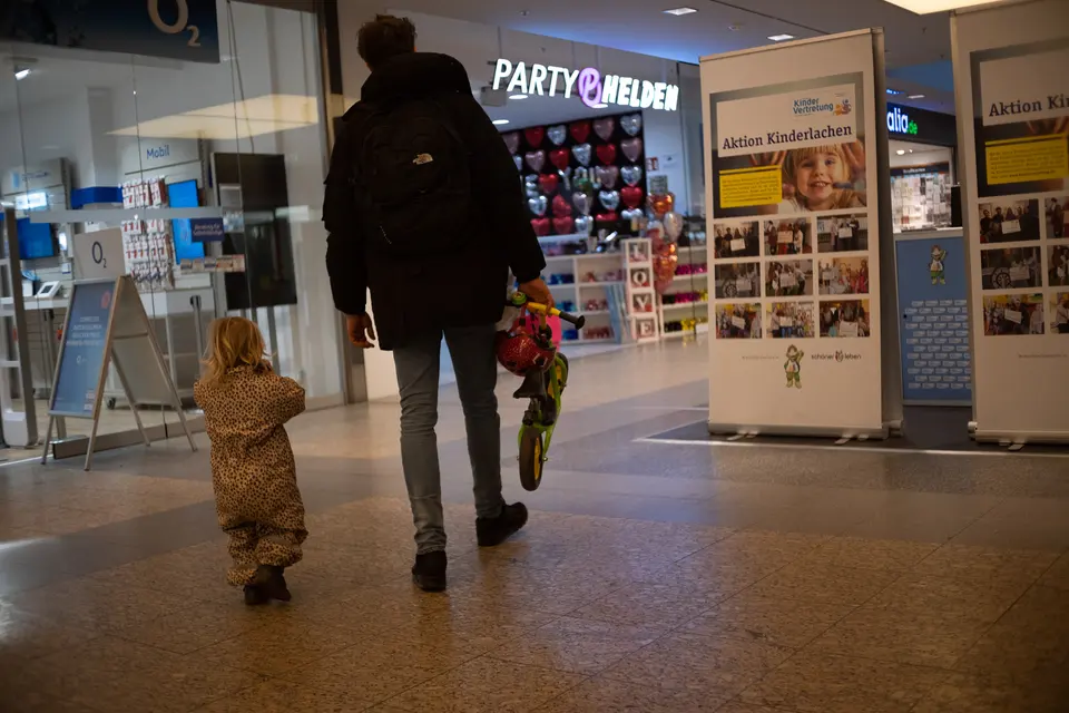 An adult and a toddler in a bear suit walking through a shopping arcade, seen from behind