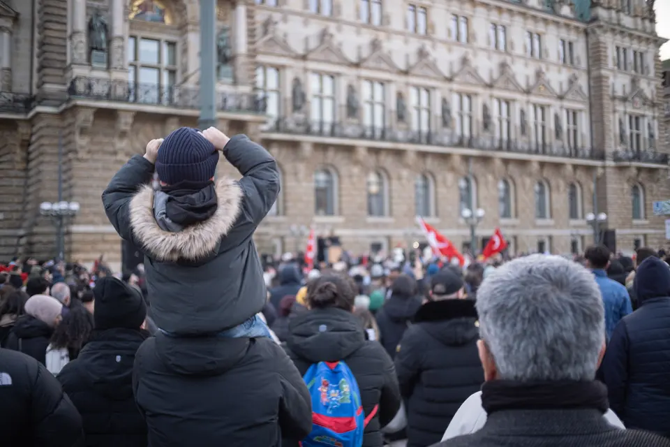 A child on an adult's shoulders, seen from behind, looking over the crowd towards the town hall
