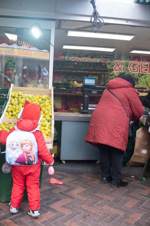 A child in a red snowsuit, seen from behind, at a greengrocer's display of fruit