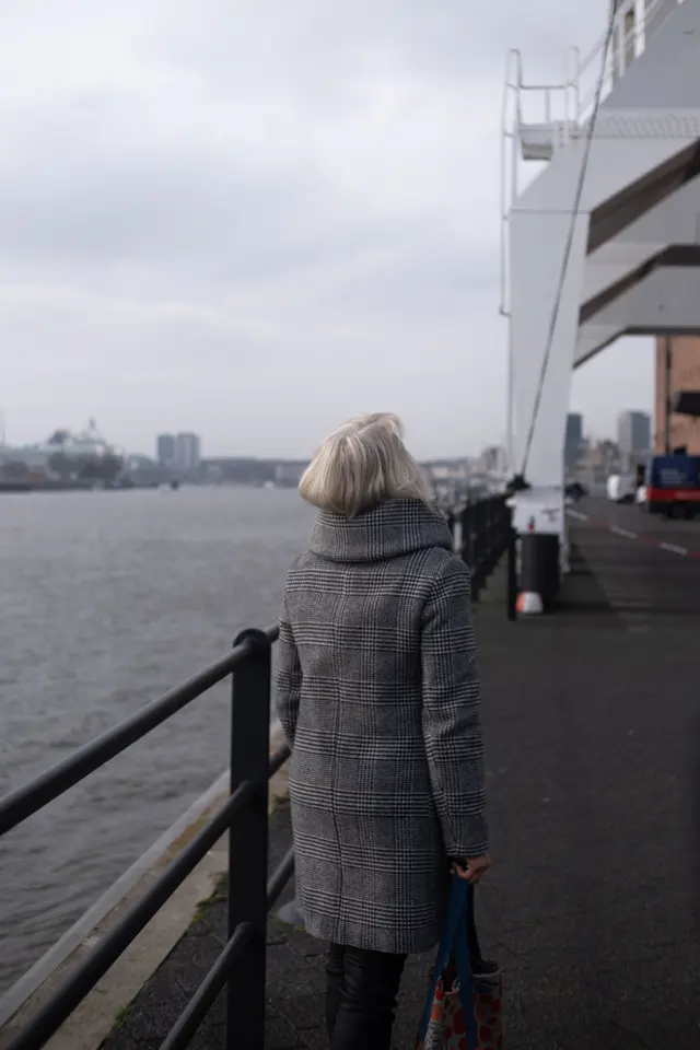 A woman in a grey coat, seen from behind, looking out over the Elbe