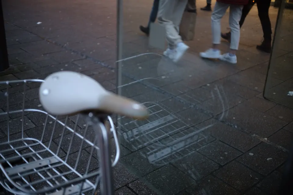 A white bicycle saddle and basket, passers-by's legs behind