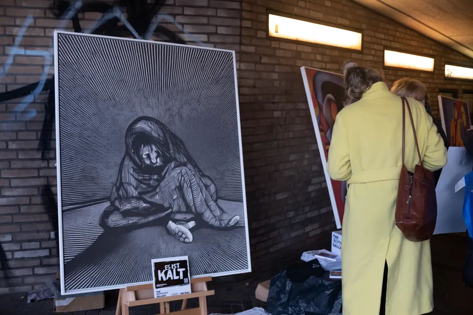 A visitor in a yellow coat looking at a black-and-white painting shown on an easel under a bridge