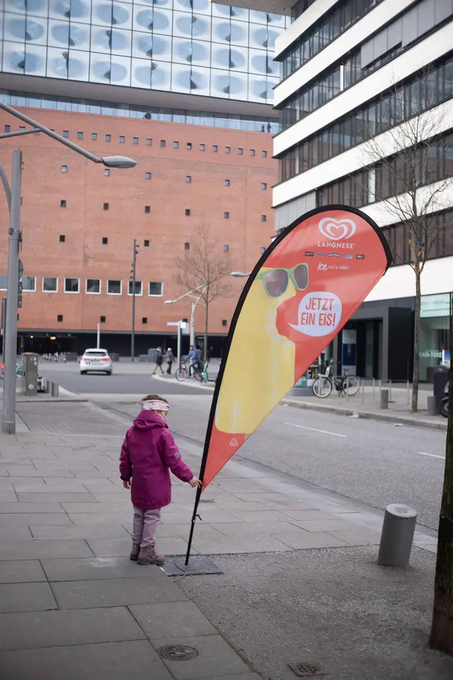 A child in a pink coat, seen from behind, beside an ice-cream flag near the Elbphilharmonie