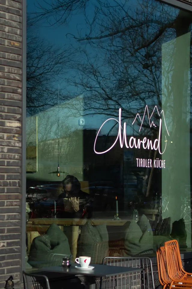 A restaurant window with a neon sign, bare trees and blue sky reflected in the glass